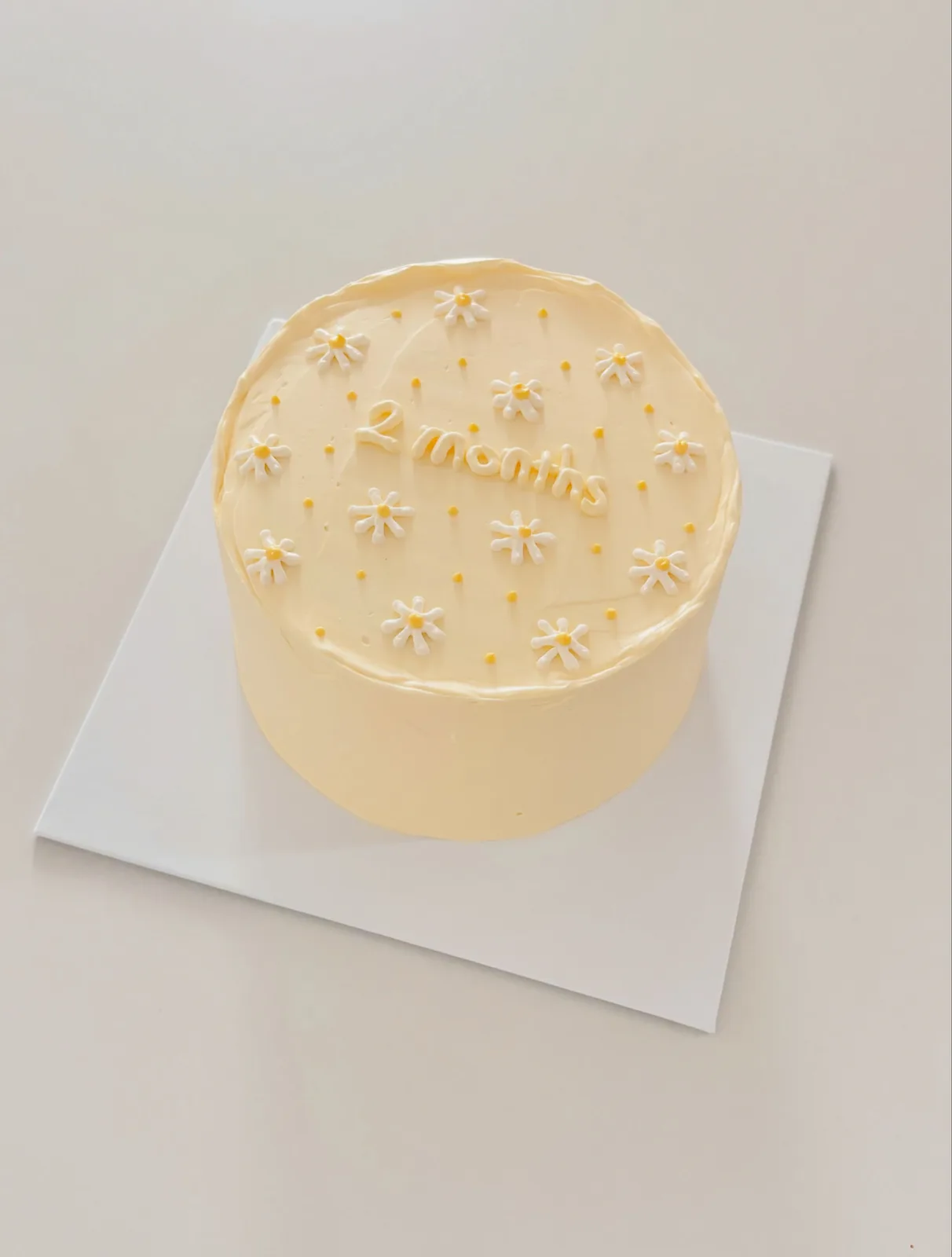 Pale yellow cake with white piped daisies and lettering, bespoke celebration cake from Danine Cakes, South London